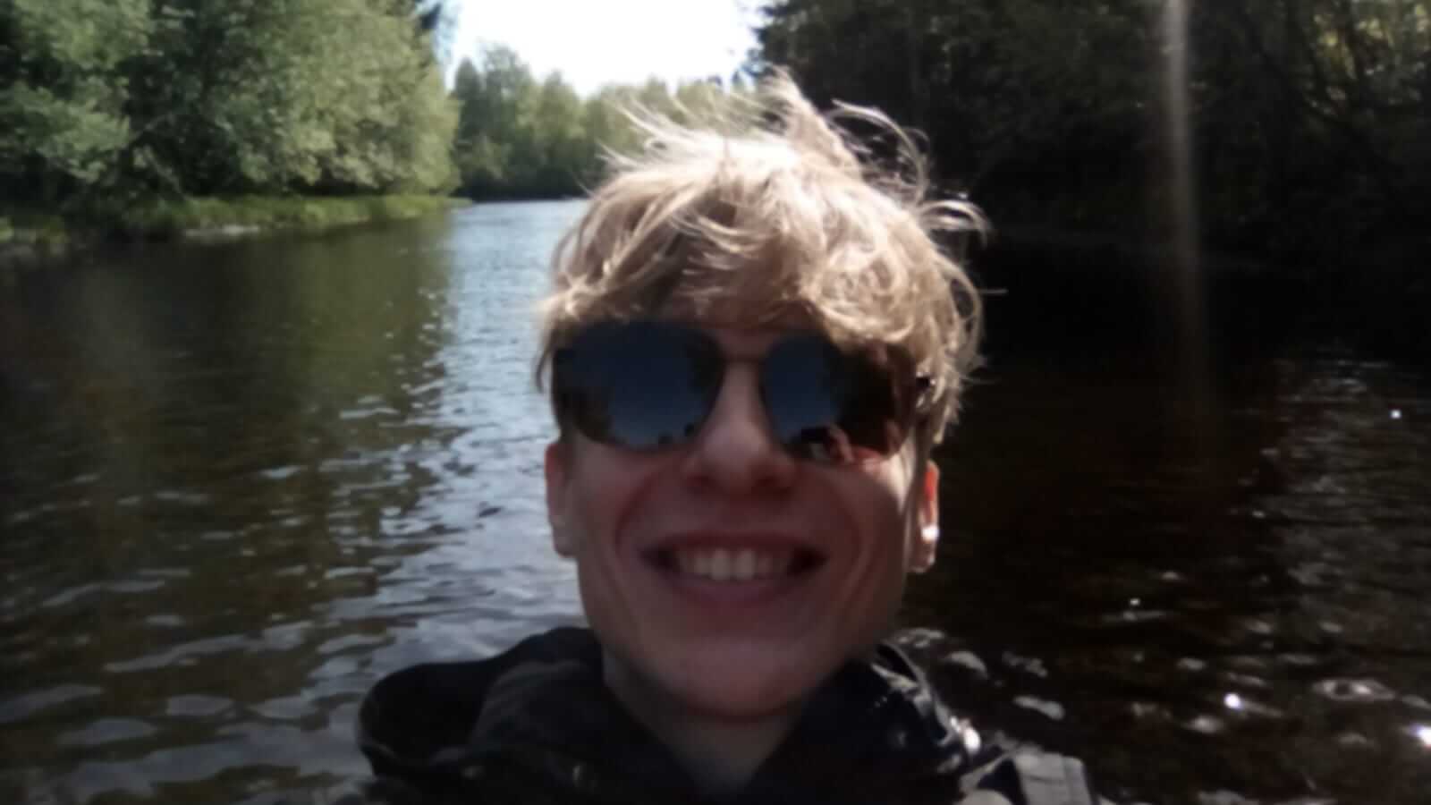 My selfie as I'm kayaking. Only my head is shown, I'm grinning widely with my teeth showing. Behind me is water and trees. My face is not focused properly which makes the photo silly.