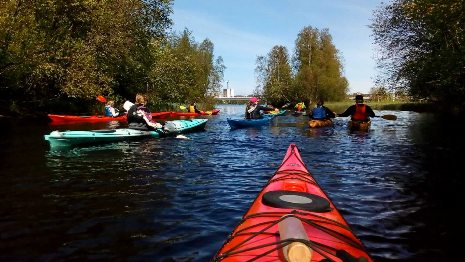 A photo taken from a red kayak which is partly showing in the bottom right corner. Eight other kayaks are in the background with people (my teammates) in them with their backs toward me. There are trees on the shores and buildings in the distant horizon.